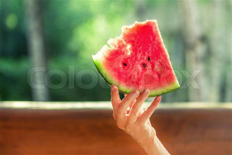 Woman holding piece of fresh tasty organic watermelon berry sitting on wooden terrace. Person on picnic outdoor with juicy ripe red melon berry in hand on bright ..., stock photo