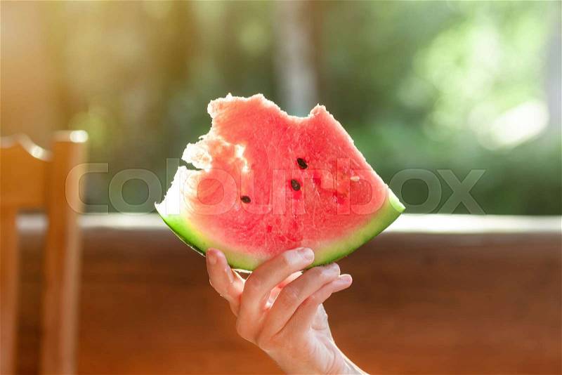 Woman holding piece of fresh tasty organic watermelon berry sitting on wooden terrace. Person on picnic outdoor with juicy ripe red melon berry in hand on bright ..., stock photo