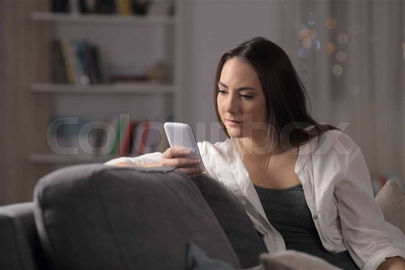 Serious woman checking smartphone content sitting on a couch in the night at home, stock photo