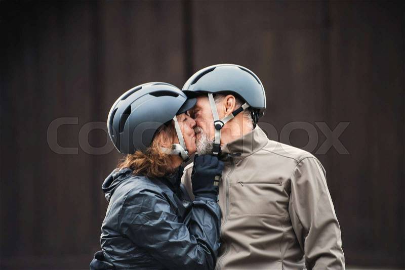 Happy active senior couple with bike helmets standing outdoors against dark background, kissing, stock photo