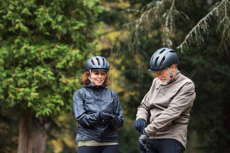 A happy senior couple with bicycle helmet standing outdoors on a road in park in autumn, stock photo