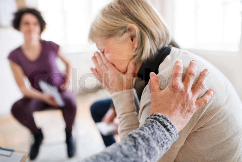 A senior depressed woman crying during group therapy, other people comforting her, stock photo