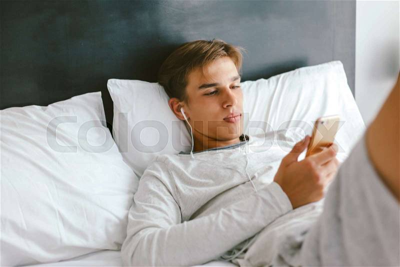 16 years old teenager using smartphone and listening to music in earphones while relaxing on couch in his room, stock photo