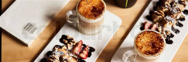 Banner size image of two cups of Hot latte with baked caramel crust and sweet roll with banana and strawberry on the wooden table in coffe-shop. Coffee concept, stock photo