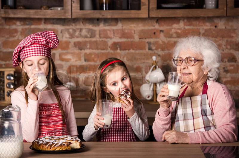 Caring granny with two little granddaughters drinking milk and tasting homemade pie, stock photo
