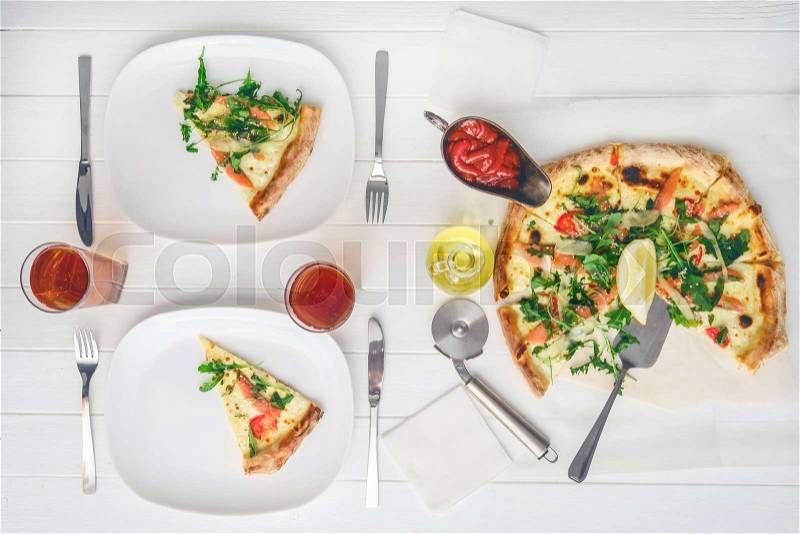Freshly cooked pizza cuts for two people with cutlery, sauces and drinks on white wood table, stock photo