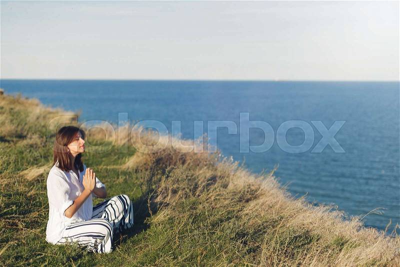 International Yoga day concept. Young beautiful woman practicing yoga on beach, sitting in grass. Hipster girl doing yoga, relaxing and listening to sea waves. ..., stock photo