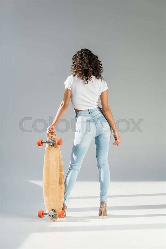Back view of woman with curly hair standing with skateboard on grey background, stock photo