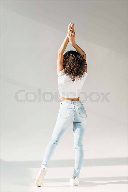 Back view of woman posing with hands above head in tight blue jeans on grey background, stock photo