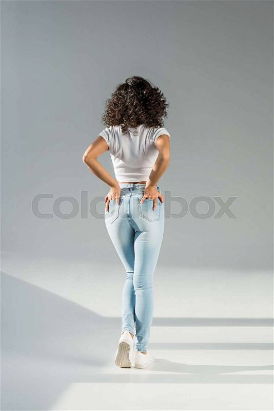Back view of woman standing with hands in pockets in tight blue jeans on grey background, stock photo