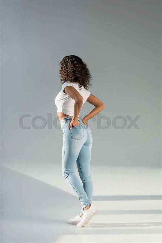 Back view of woman standing with hands in pockets in sexy blue jeans on grey background, stock photo