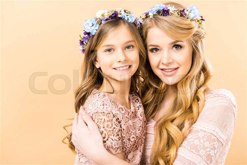 Cute child and pretty mom in colorful floral wreaths embracing and looking at camera on yellow background, stock photo