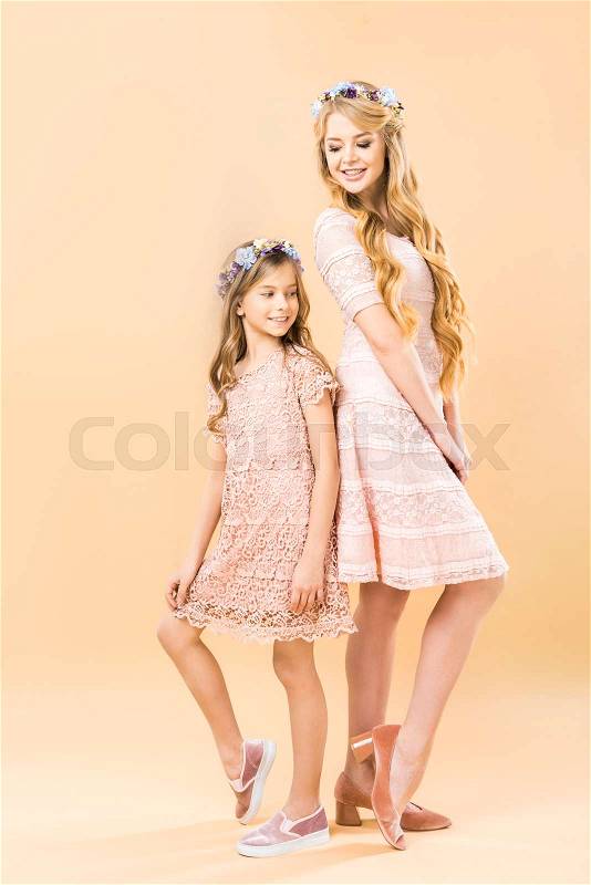 Mom and daughter in elegant lacy dresses and floral wreaths standing back to back and posing at camera on yellow background, stock photo