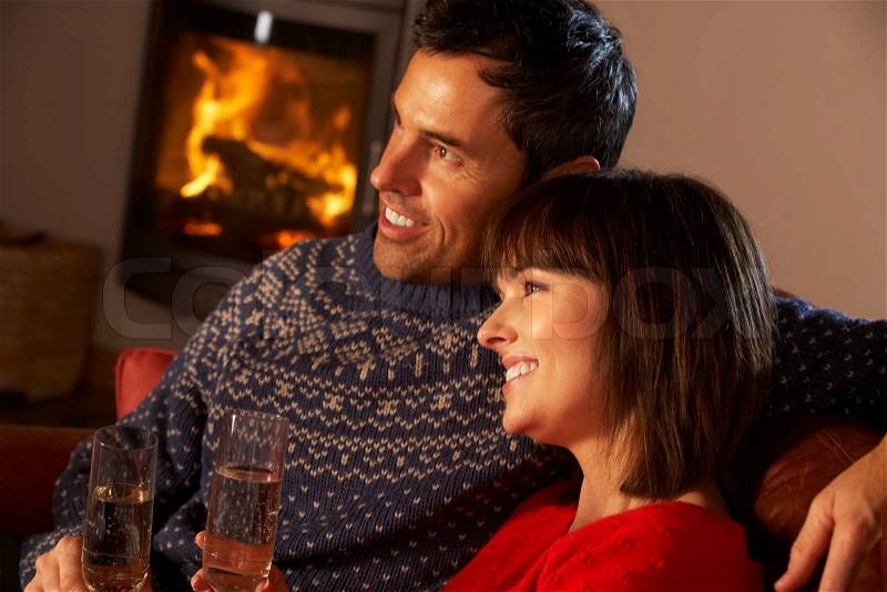 Middle Aged Couple Sitting Sofa By Cosy Log Fire With Glass Of Champagne, stock photo