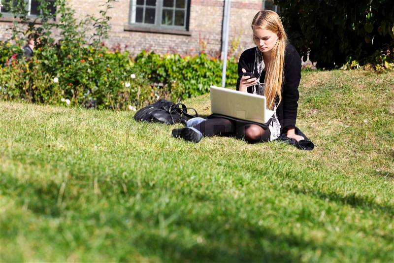Young blond student sitting at campus lawn with apple phone and notebook, stock photo