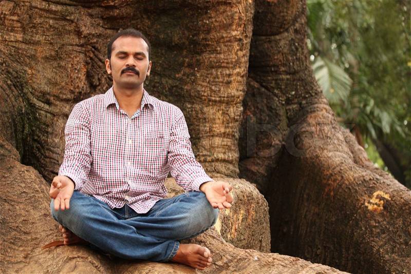 Close-up photo of a handsome indian executive doing meditation under a tree in a park The person is sitting in lotus posture and is calm, relaxed closing his eyes for better concentration, stock photo