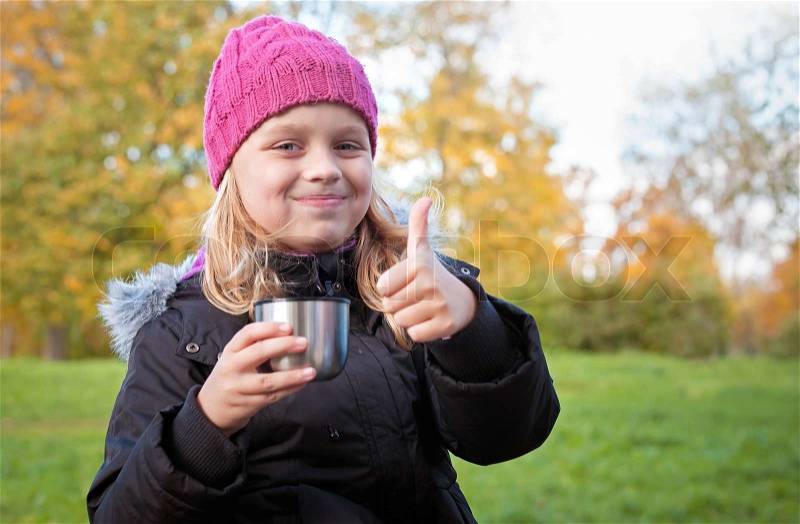 Little beautiful blond girl drinks tea in autumnal park and showing thumbs up, stock photo