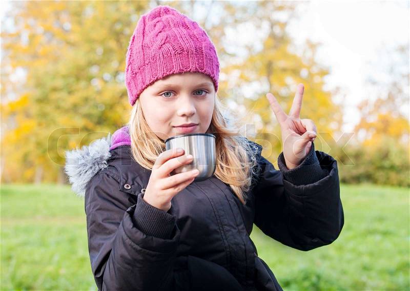 Little beautiful blond girl drinks tea in autumnal park and showing victory fingers, stock photo
