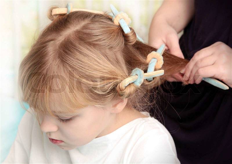 Little blond Russian girl with curlers on hair and hairdressers hands, stock photo