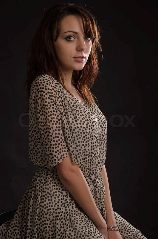 Beautiful young brunette woman posing sideways looking at the camera in a stylish summer dress with dots against a dark studio background, stock photo