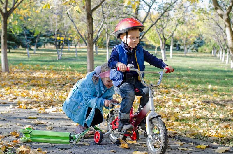 Cute little boy learning to ride his new bicycle fitted with training wheels helped by his young sister as the two play together in the park, stock photo