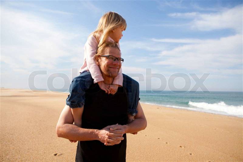 Father carrying young daughter piggyback on shoulders at beach, stock photo