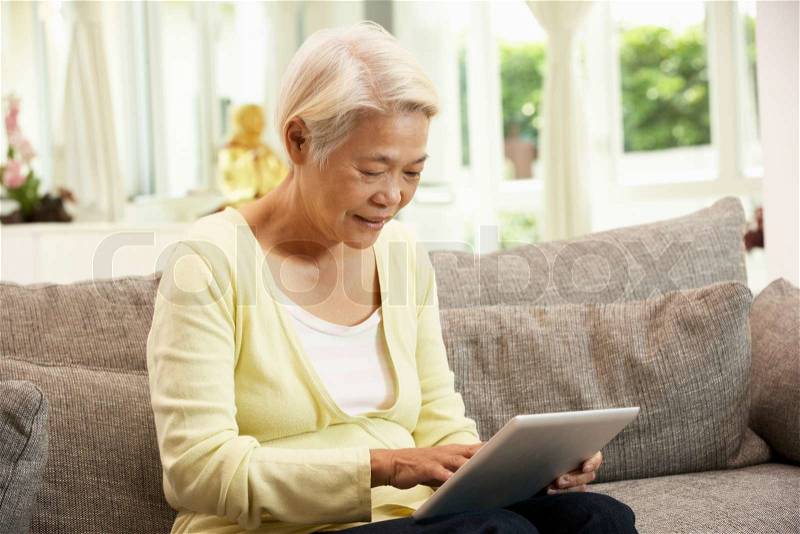 Senior Chinese Woman With Tablet Computer Whilst Relaxing On Sofa At Home, stock photo