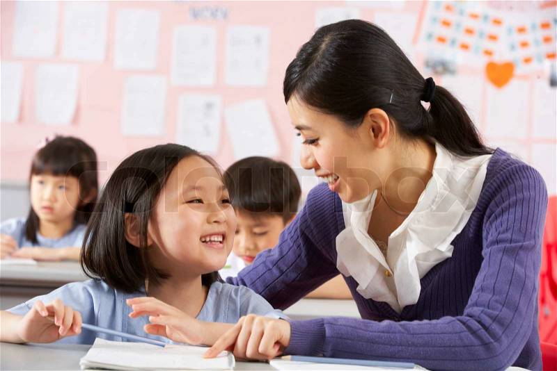Teacher Helping Student Working At Desk In Chinese School Classroom, stock photo