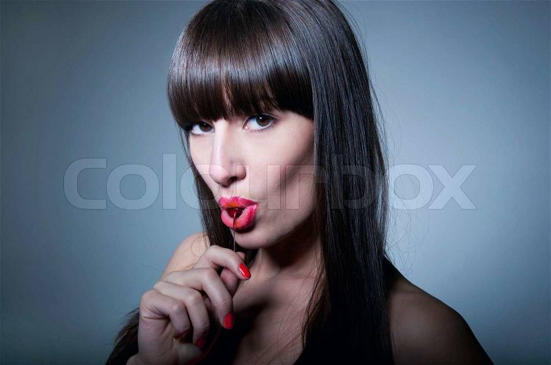 Glamour portrait of sexy nude woman model with natural makeup, long healthy shiny hair and long fringe, licking ripe fresh cherry and looking at camera with temptation. Gray background, stock photo