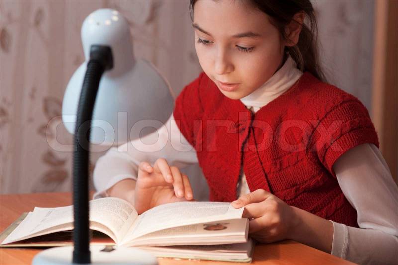 The girl at the desk reading a book by the light of the lamp, stock photo