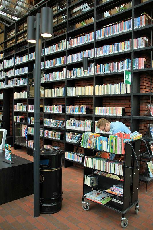 The boy, student, is working between all kind of books in different colours in the municipal library, stock photo