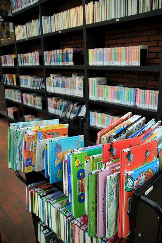 Children\'s books on a cart and a bookcase with all kind of books in different colours to hire in the library, stock photo
