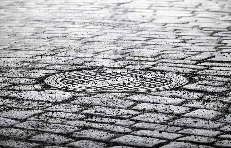 Round steel sewer manhole on dark old cobblestone road, stock photo