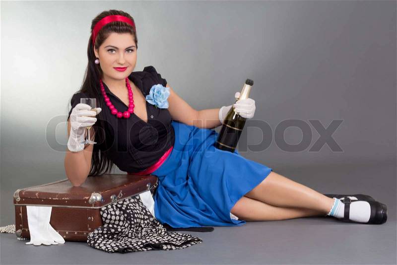 Pinup girl with bottle of champagne and packed suitcase over grey, stock photo