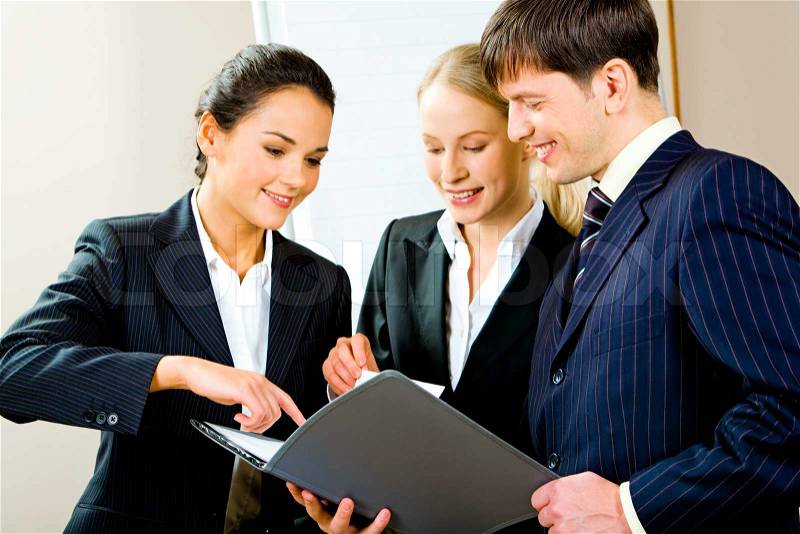 Portrait of three smiling young specialists looking at business plan with one of businesswomen pointing at something in it, stock photo