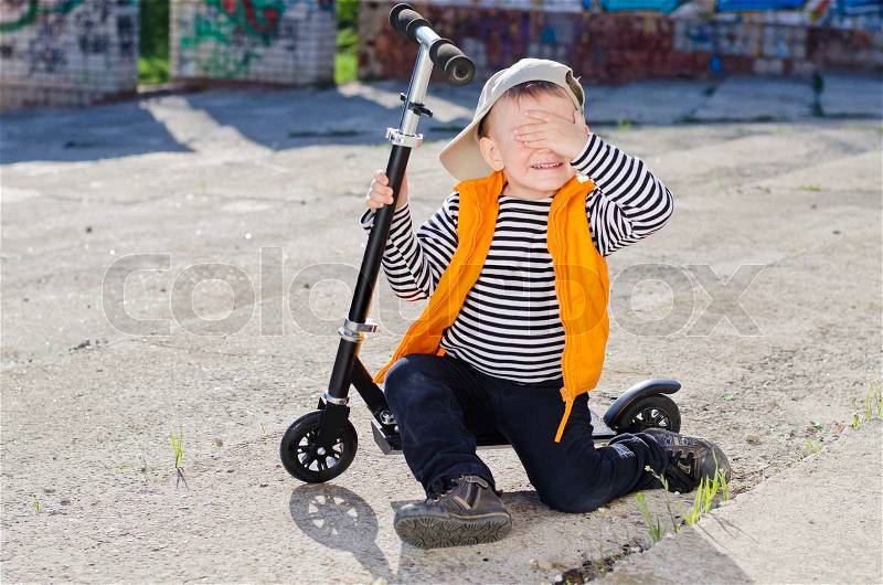 Bashful little boy sitting on the footboard of his scooter covering his eyes from the camera, stock photo