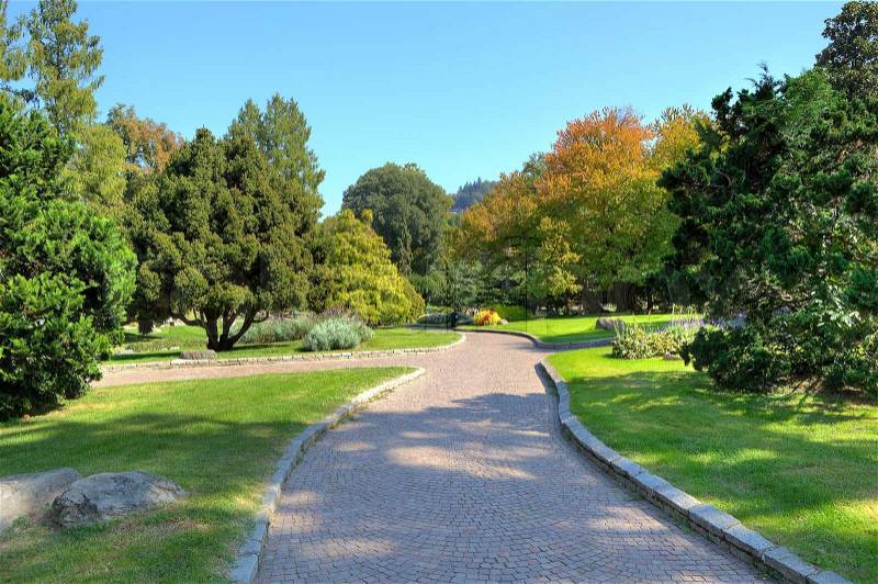 Narrow cobbled alley among green lawns and trees under clear blue sky at botanical part of famous Valentino Park in Turin, Italy, stock photo