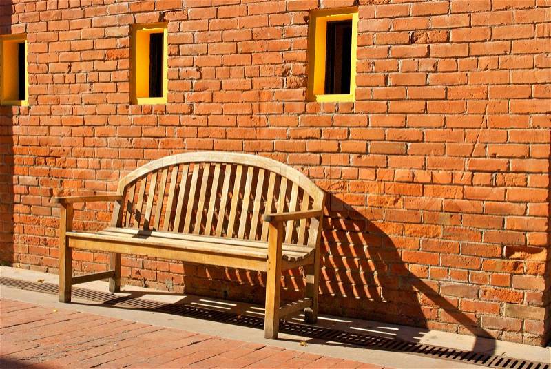 An old wooden bench sits by a worn orange brick wall with three yellow windows, stock photo