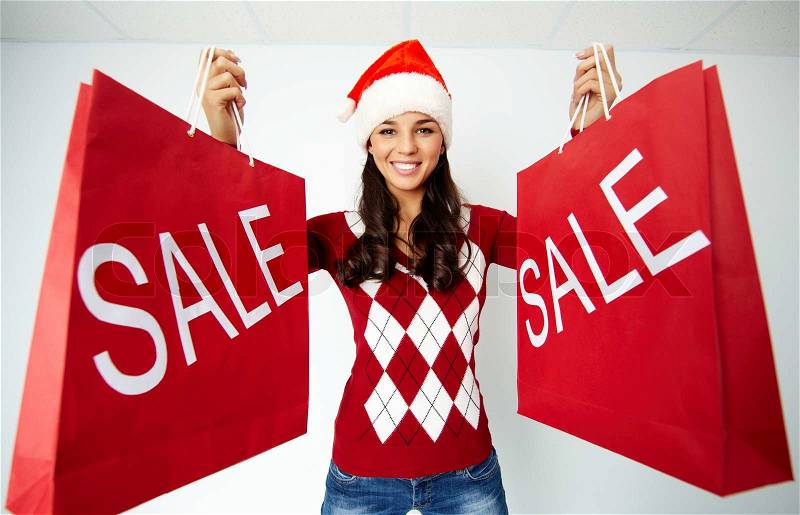 Happy girl in Santa cap holds red paperbags with symbol of sale, stock photo