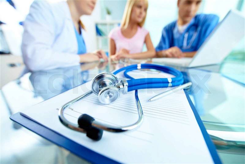 Close-up of stethoscope and paper on background of doctors and patient working with laptop, stock photo