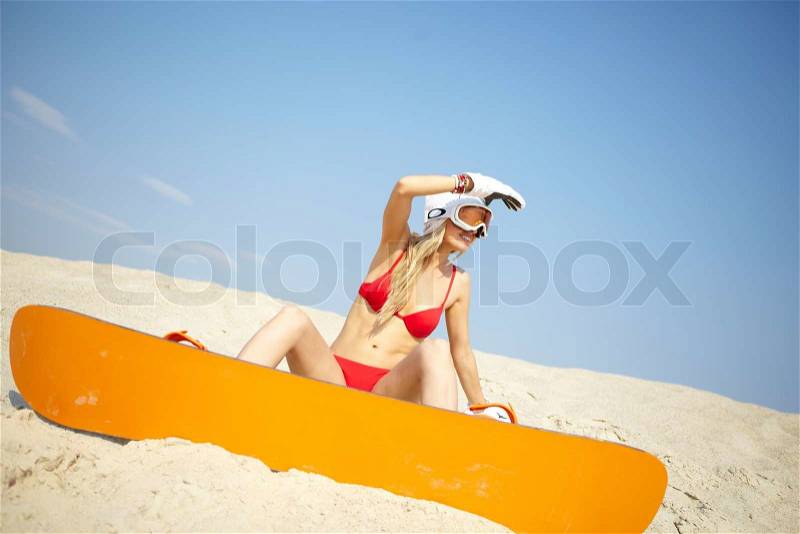 Young blonde woman sitting on sand with a snowboard and looking around, stock photo
