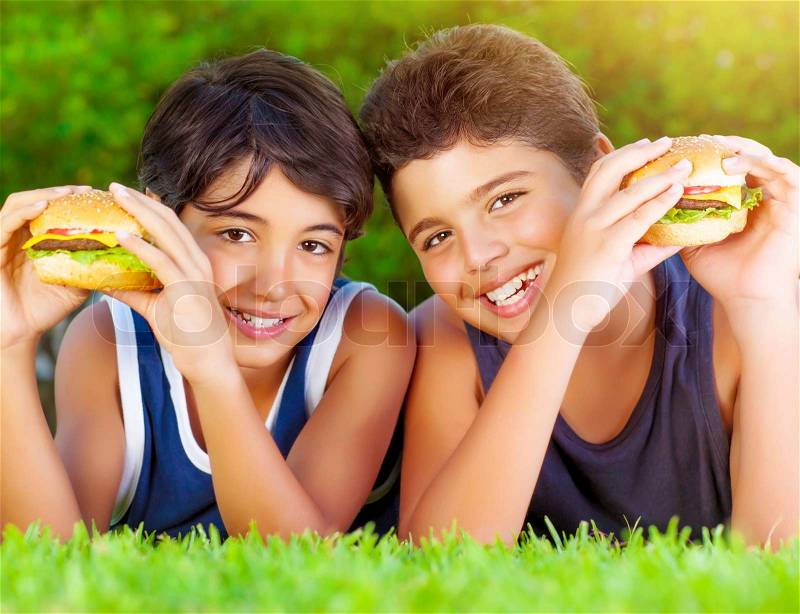 Closeup portrait of two happy boys eating big tasty fatty burgers outdoors, lying down on green field and enjoying sandwich with cheese, meat and vegetables, stock photo