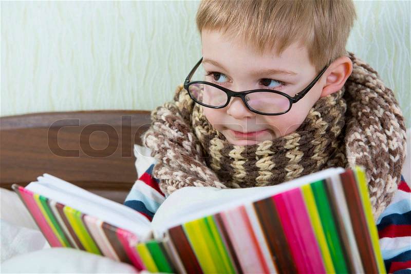 Little sick boy in glasses reading book in bed, stock photo