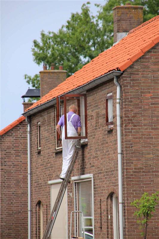 The painter in purple coverall stands on the stairs is painting the window frame in summertime, stock photo