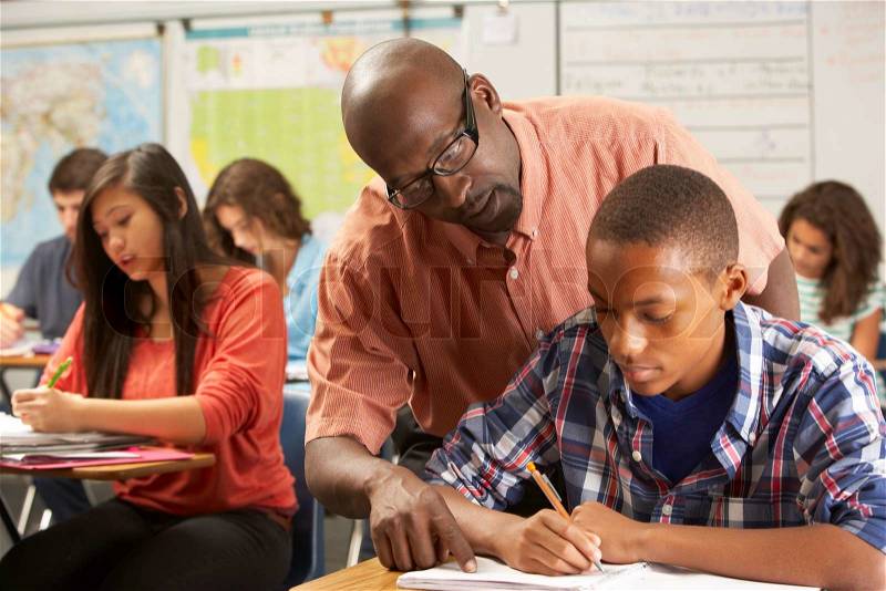 Teacher Helping Male Pupil Studying At Desk In Classroom, stock photo