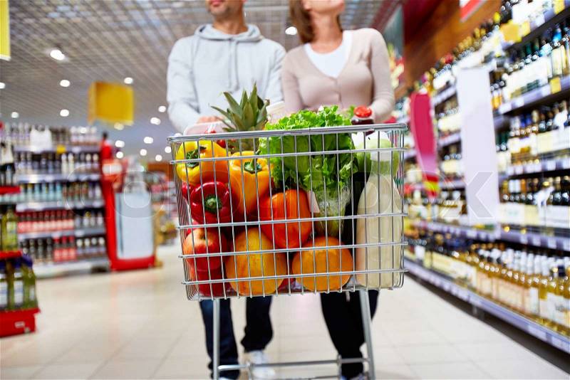 Image of cart full of products in supermarket being pushed by couple, stock photo