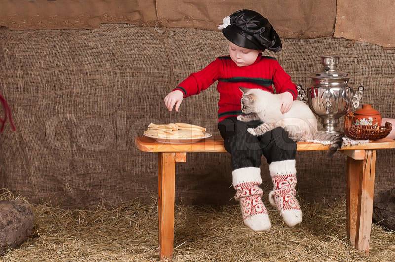 Boy in national costume sits in the cart, there are pancakes and plays with a cat sitting on the bench, stock photo