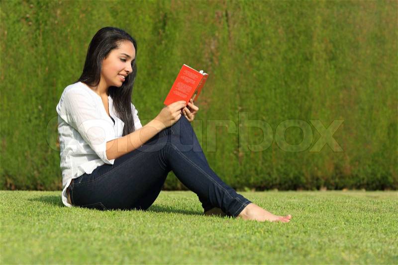 Beautiful arab woman reading a book sitting on the lawn of a park with a green background, stock photo