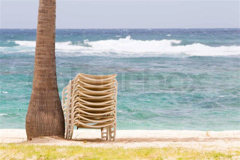 Stack of beach chairs under palm tree on idyllic tropical white sand beach, stock photo