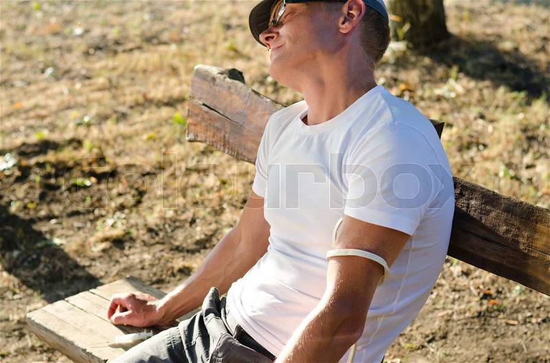 Addicted Caucasian man sitting on a bench in the park feeling side effects after injecting a drug dose intravenously, stock photo